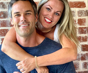 Rory McKernan stands in front of a brick wall with his wife hugging him from behind
