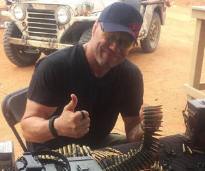 Randy Couture gives a thumbs up and holds up a string of bullets with a tan jeep in the background
